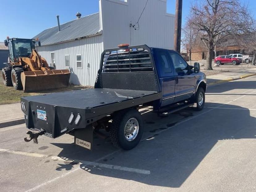 Blue truck with flatbed parked on street, featuring a metal rear bumper and visible construction equipment nearby.